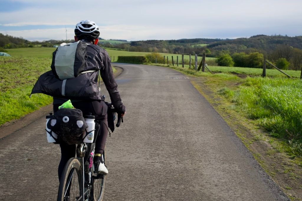 Cycliste vu de dos sur une petite route de campagne, coupe-vent noir gonflé par l’air sur le dos.