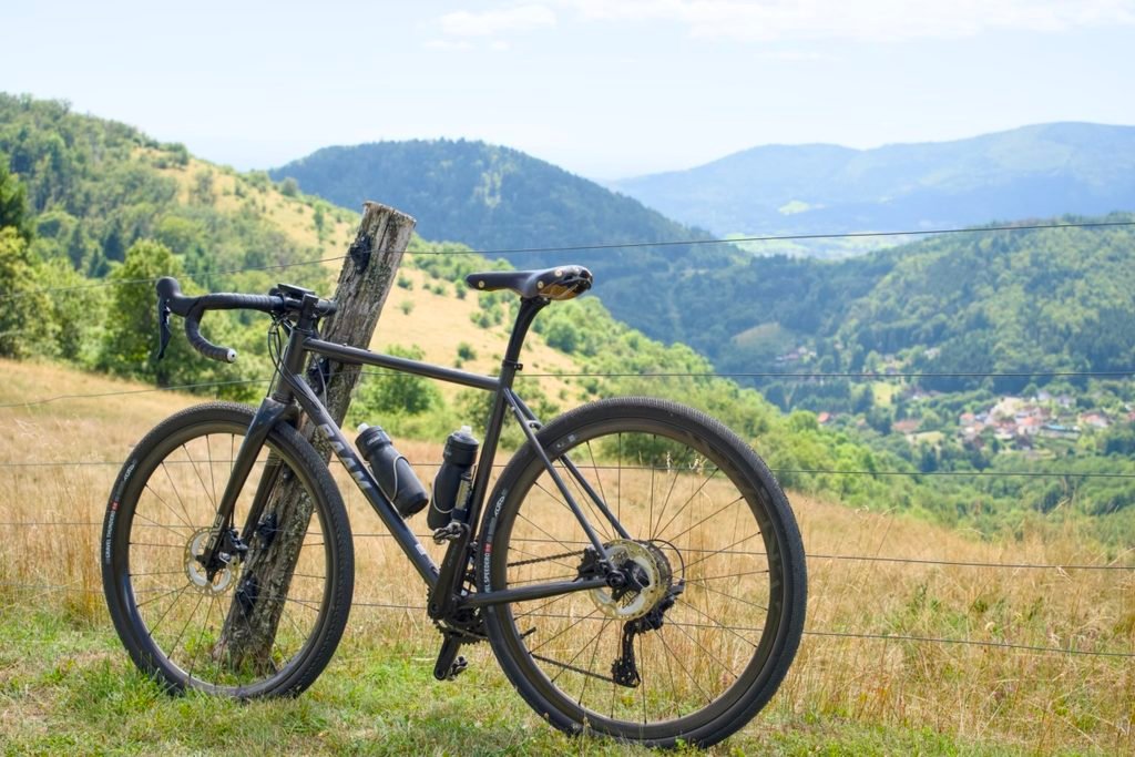 Gravel acier : ce que plus de 2 ans sur un Reynolds 853 m'ont appris 8 Gravel acier BAAM Argh appuyé contre un piquet de clôture dans une pâture de montagne, avec une vallée et un village en contrebas.