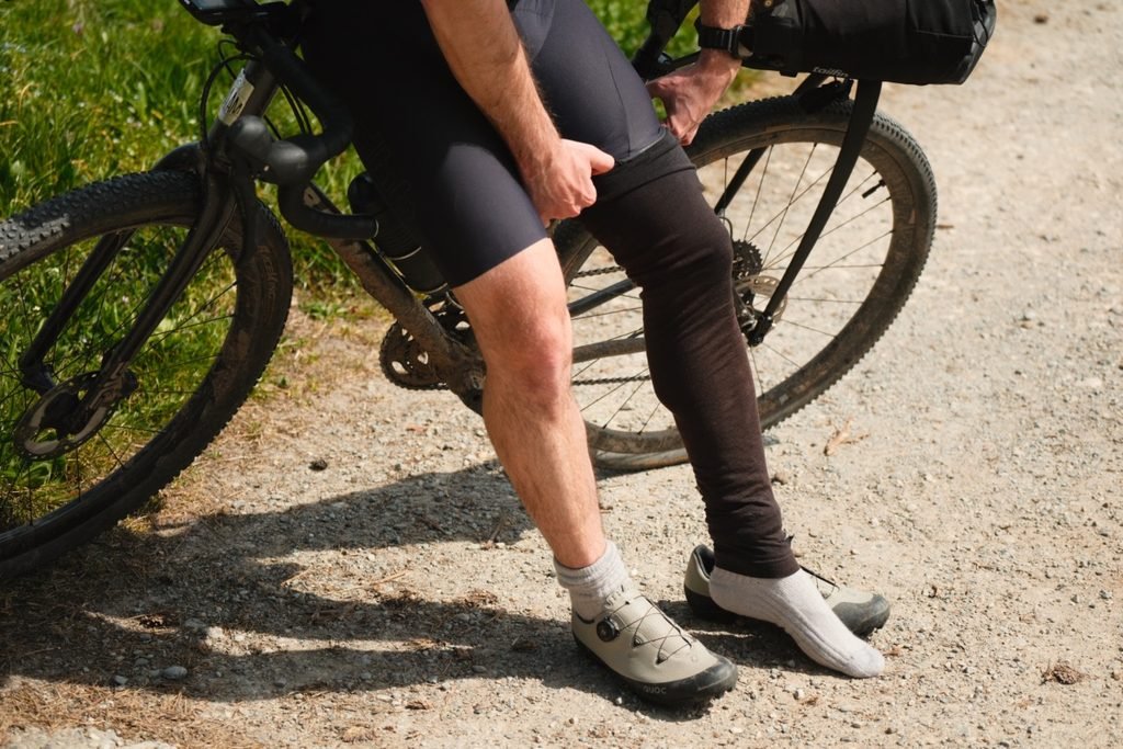 Cycliste gravel en cuissard court qui ajuste une jambière en mérinos au bord d’un chemin, l’autre jambe encore nue, chaussures Quoc aux pieds.
