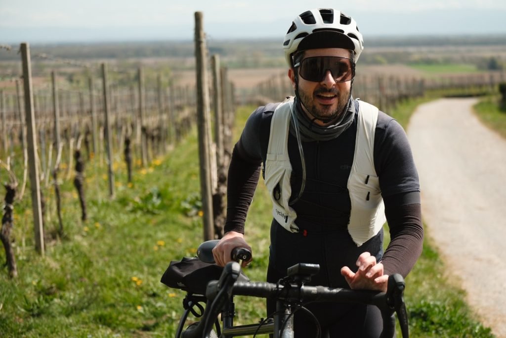 Cycliste en maillot gravel à manches courtes avec manchettes et tour de cou, gilet d’hydratation sur les épaules, à côté de son vélo dans les vignes.