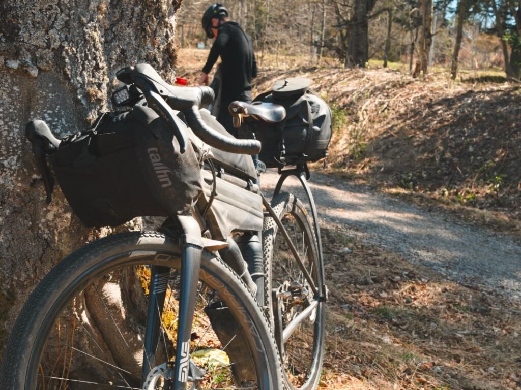 Quelle sacoche gravel pour rouler à la journée ? 5 Gravel chargé appuyé contre un arbre, avec une sacoche de guidon Tailfin, une sacoche de cadre et une sacoche arrière, au bord d’un chemin
