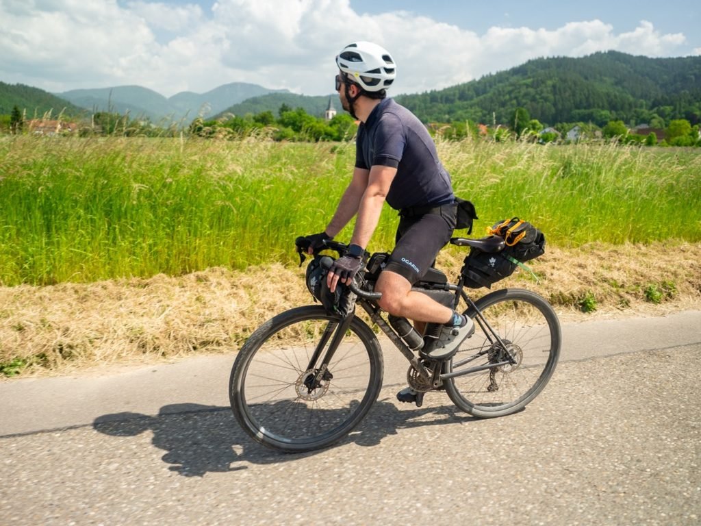 Cycliste en tenue gravel d’été, maillot et cuissard courts avec gants courts, sur une petite route au pied de collines boisées en plein soleil.