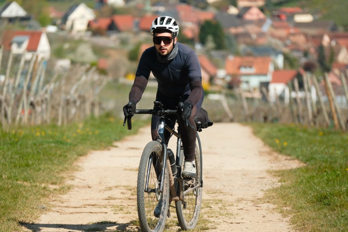 Cycliste en tenue gravel de mi-saison, maillot court avec manchettes, jambières, gants longs et tour de cou, sur un chemin de terre entre les vignes.